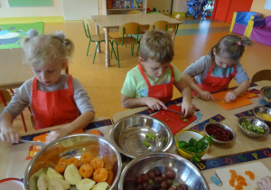 Troje dzieci siedzi przy stole, w ręku trzymają noże, którymi kroją owoce na desce. Na środku stołu stoją miski wypełnione owocami.